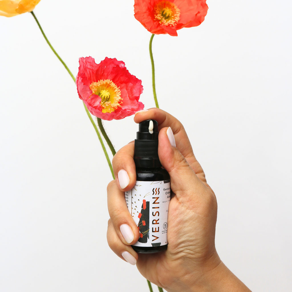 Hand holding a bottle labeled 'VERSIN' with red flowers in the background