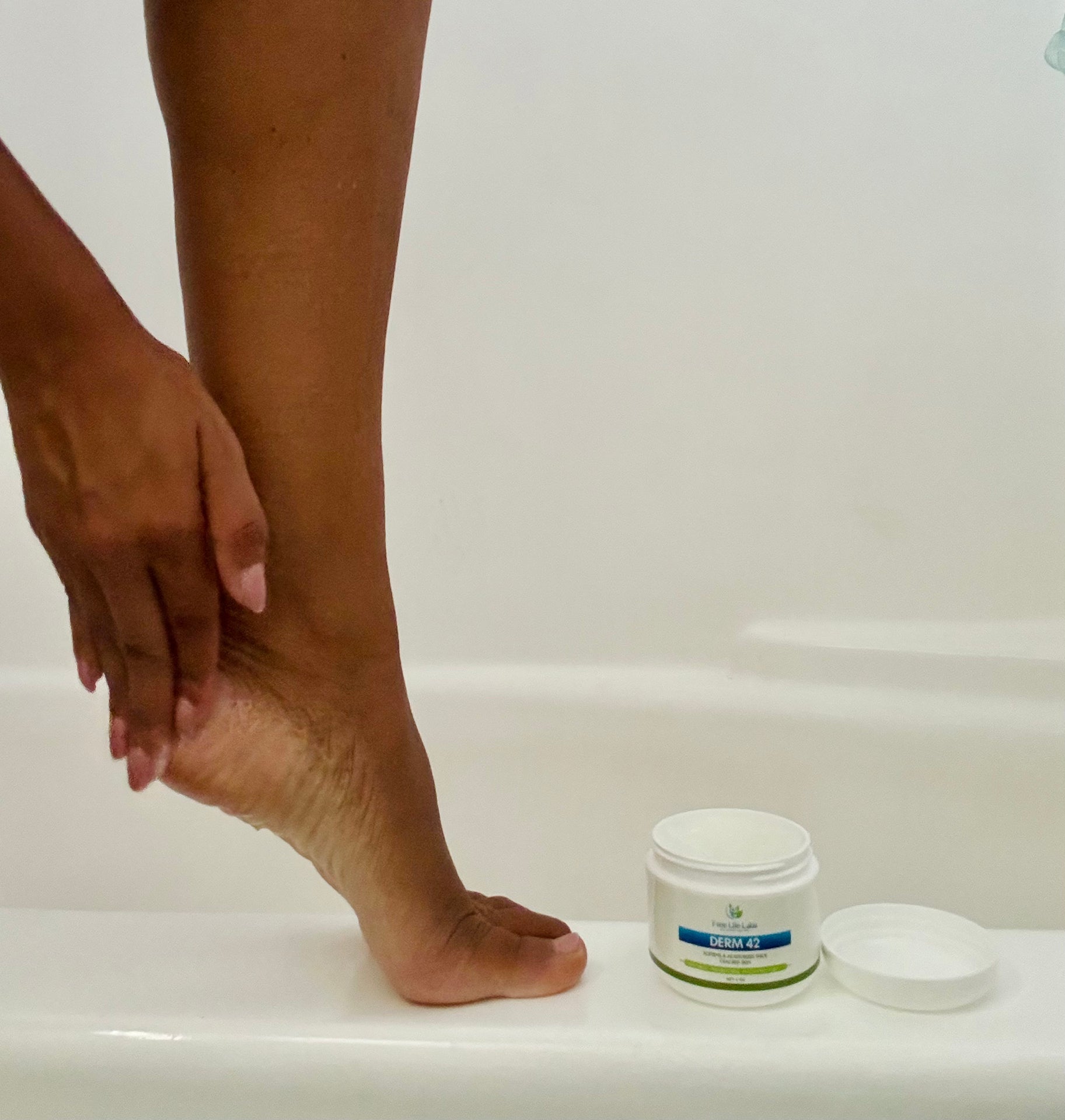 Person applying a cream to their foot next to a jar of the same cream on a white surface.