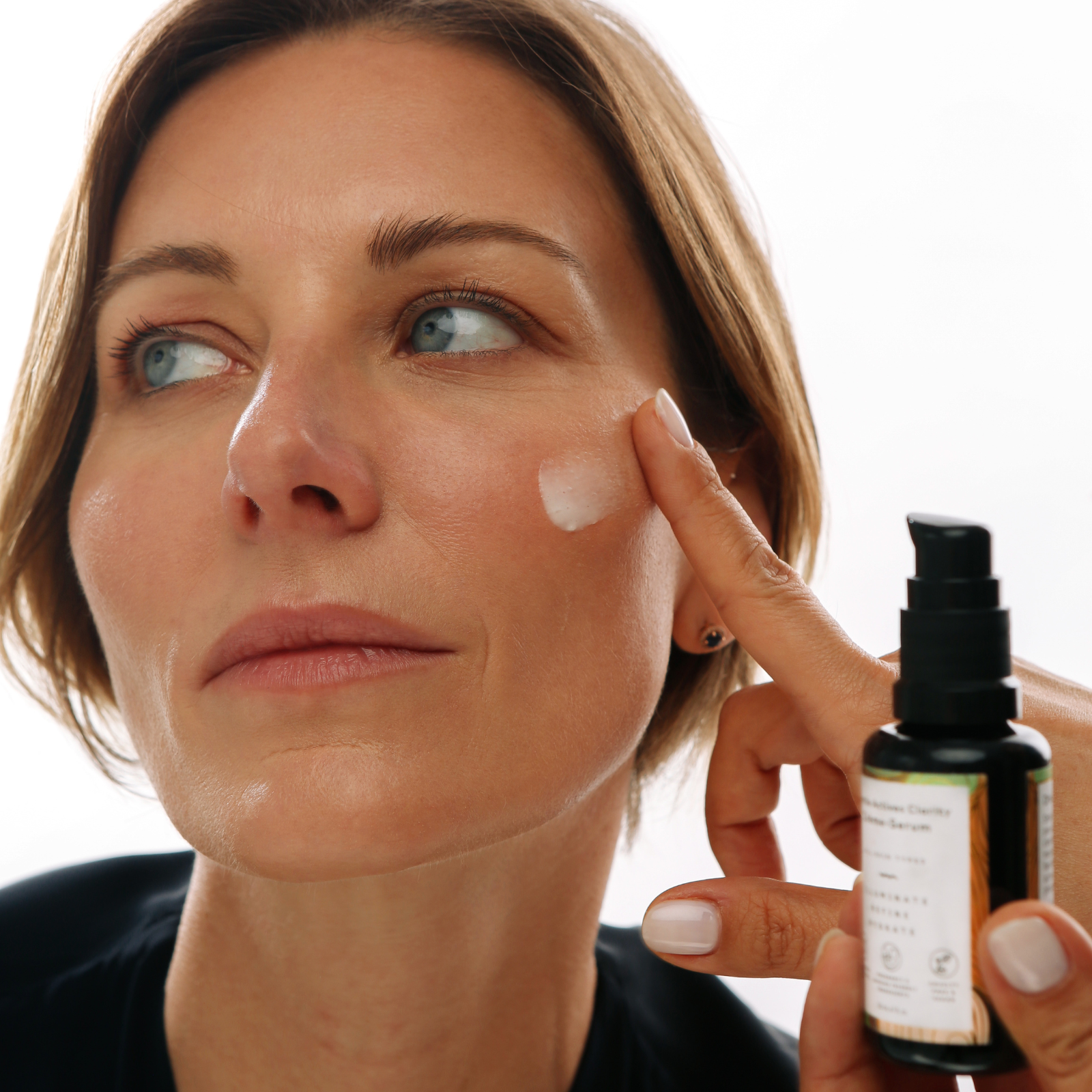 Woman applying cream to her face with a bottle of skincare product in hand.