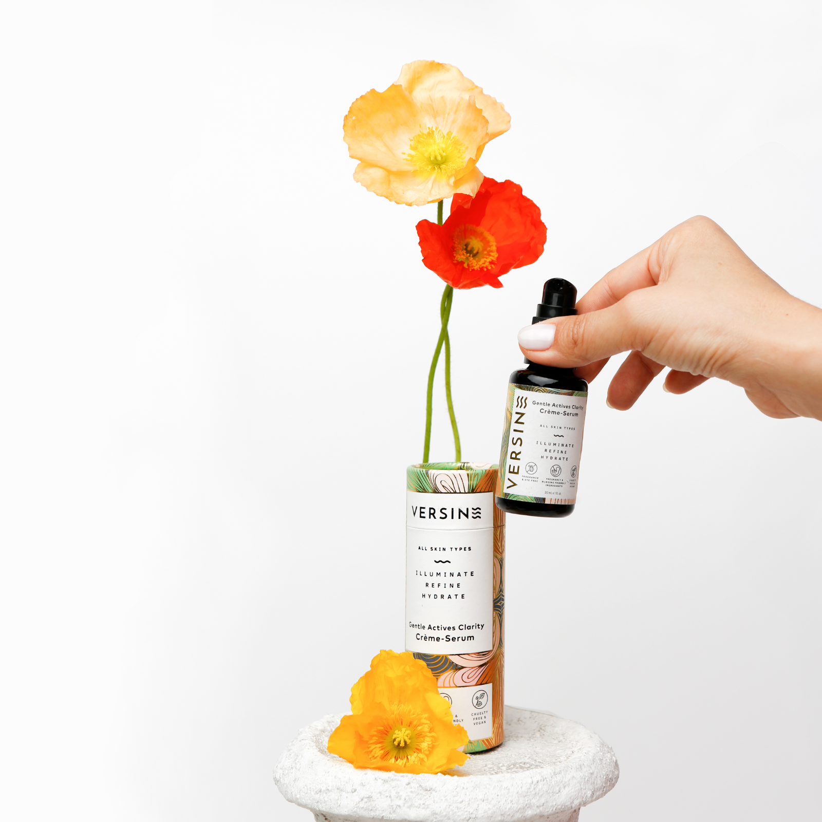 Hand holding a dropper bottle with Versine skincare product next to flowers on a white background