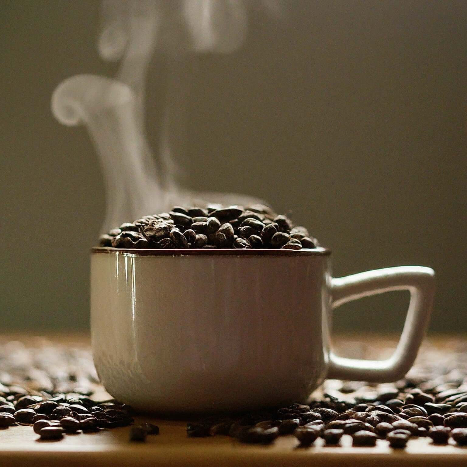 White mug filled with coffee beans on a wooden surface with steam rising.