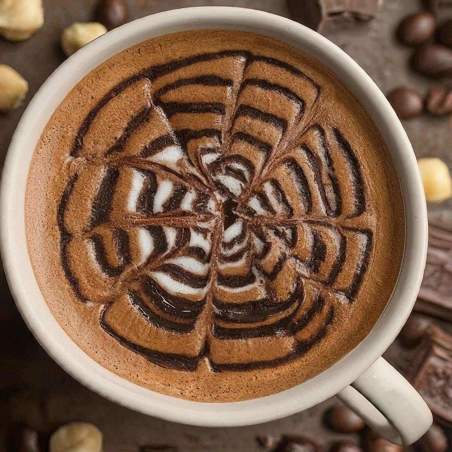 Cup of coffee with intricate latte art surrounded by coffee beans and hazelnuts.