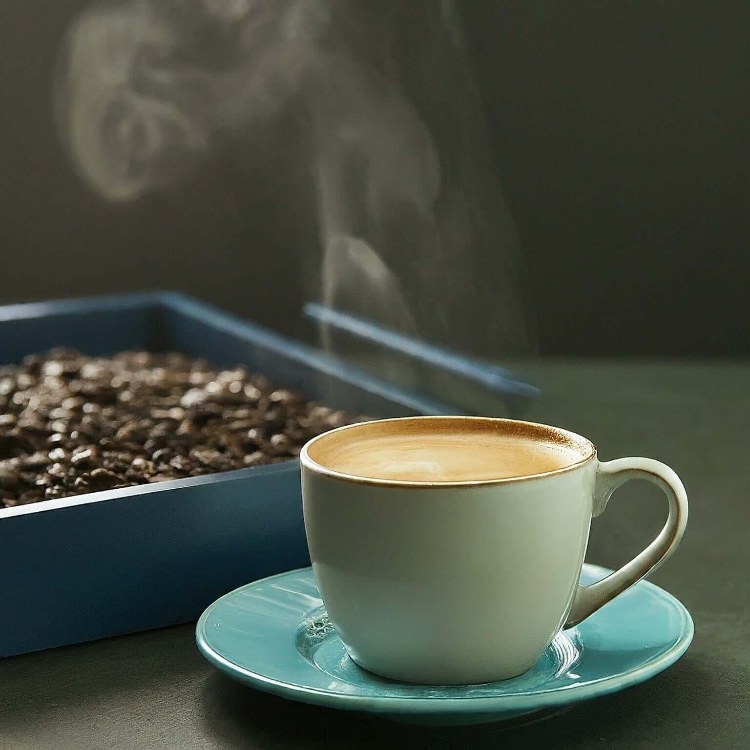 Steaming cup of coffee on a saucer with a tray of coffee beans in the background.