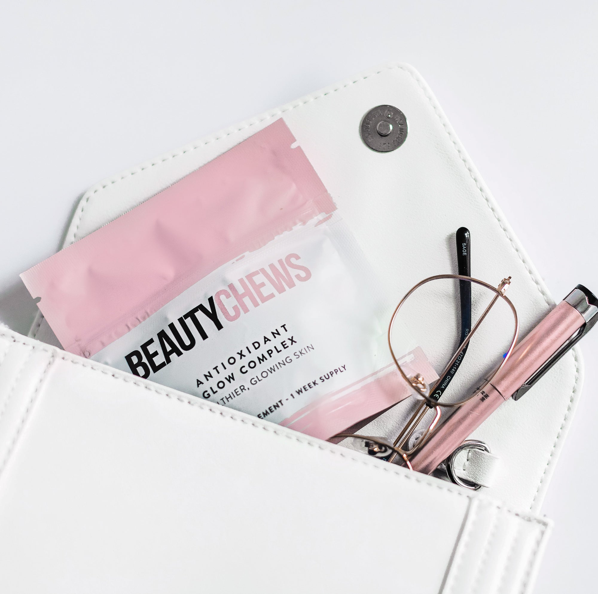 White handbag with pink and clear 'BeautyChews' package, glasses, and pen on a white background