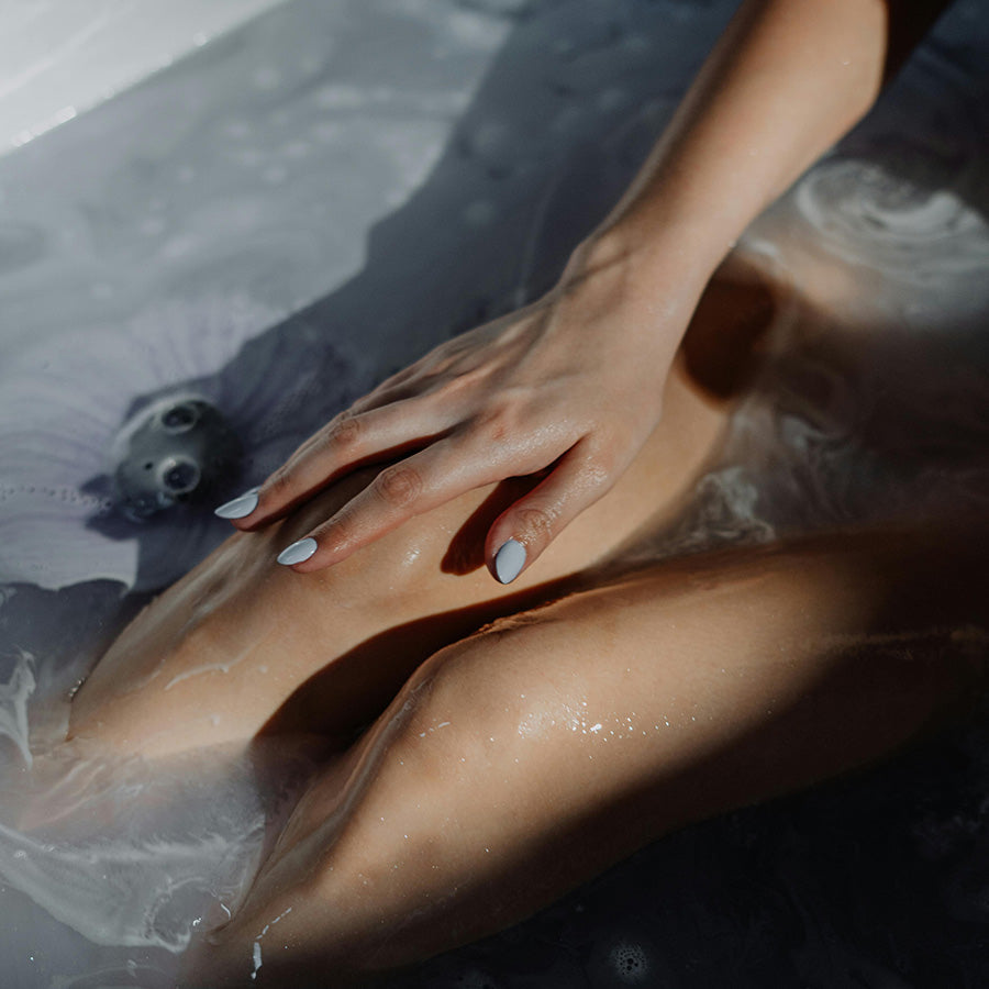 Close-up of a person's hand and leg in a bathtub with water and soap bubbles.